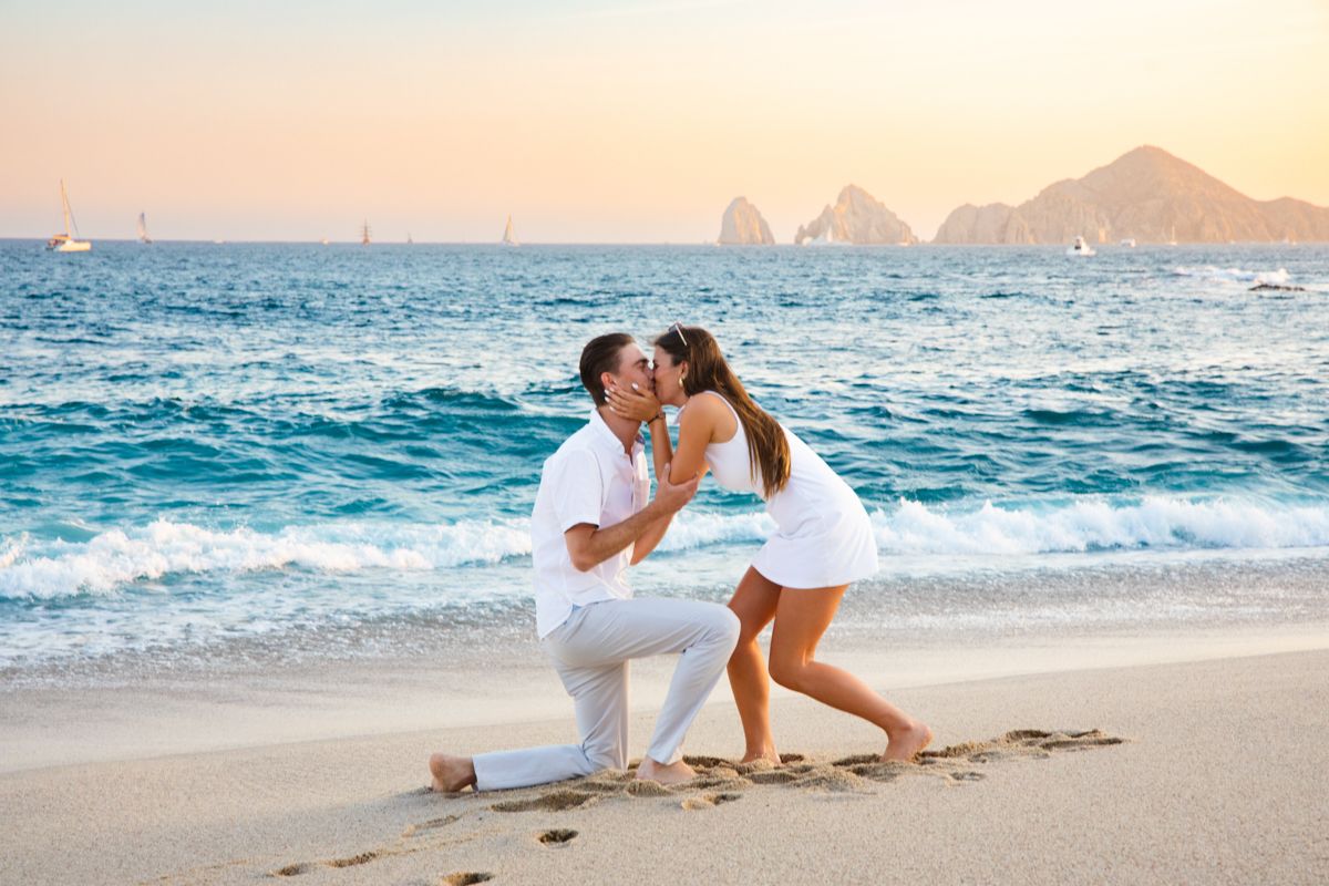 Couple sharing first kiss after beach proposal at sunset in Cabo San Lucas with ocean backdrop.
