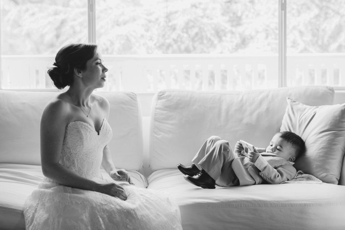 Bride in white gown sits on sofa with baby in black and white wedding photo.