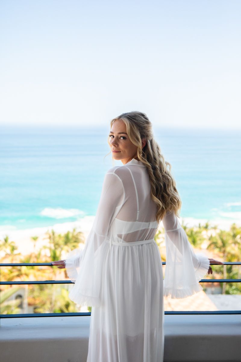 Bride in white sheer bell-sleeve robe stands on balcony overlooking turquoise ocean.
