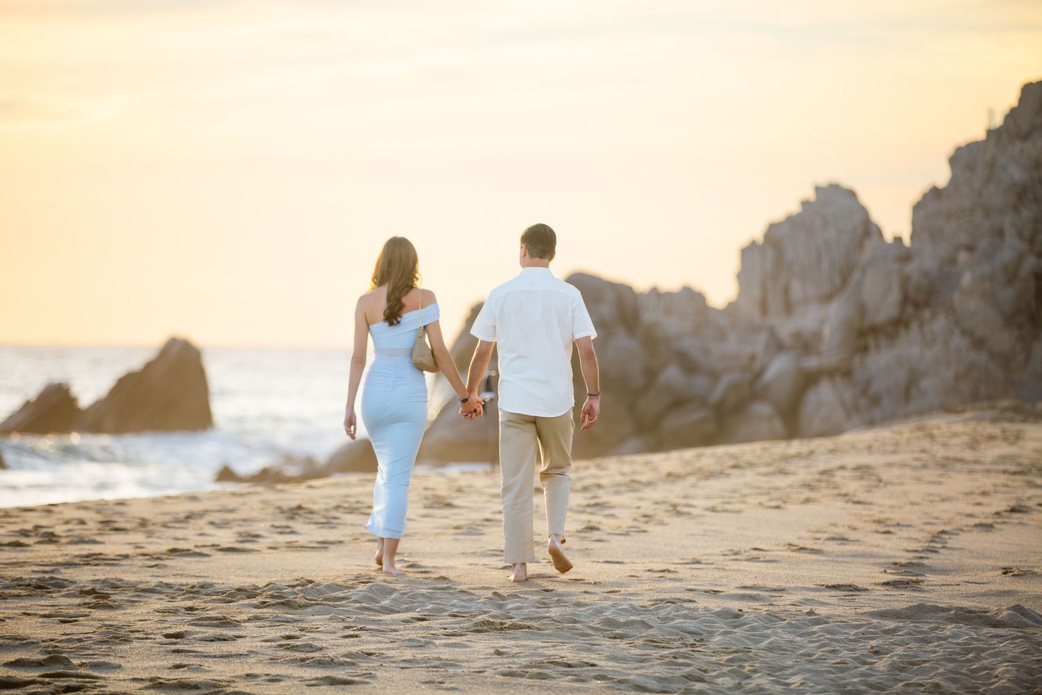 Couple holding hands walking on sandy beach  in cabo at sunset with rocky cliffs in the background
