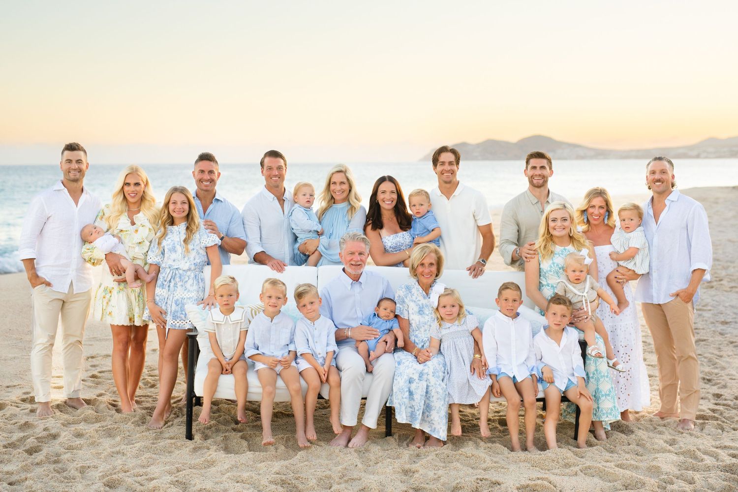 Large extended family posing together on a sandy beach in cabo at sunset in coordinating white and blue outfits.