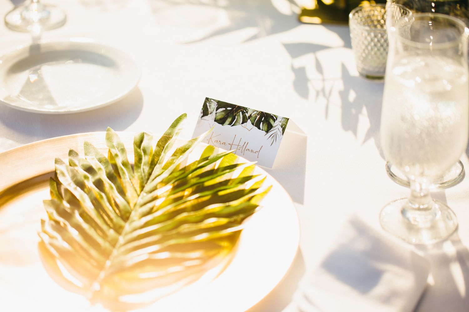 Gold palm leaf place card holder on white plate at elegant tropical wedding table setting.