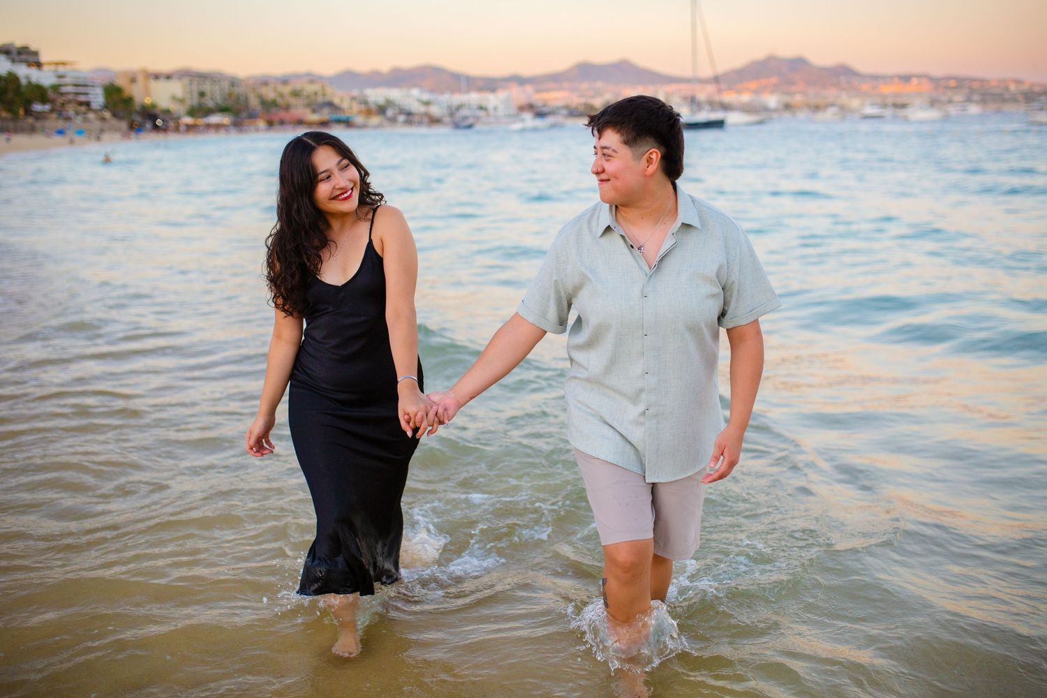 Couple holding hands walking in shallow ocean water at sunset on a scenic beach with mountains in the background in Cabo San Lucas.