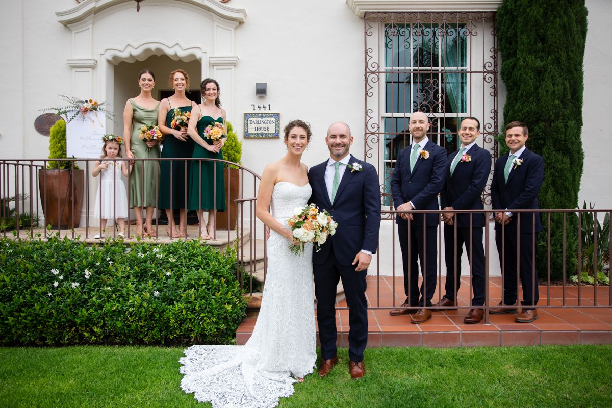 Wedding party poses outside white Spanish-style building, bridesmaids in green dresses, groomsmen in navy suits.