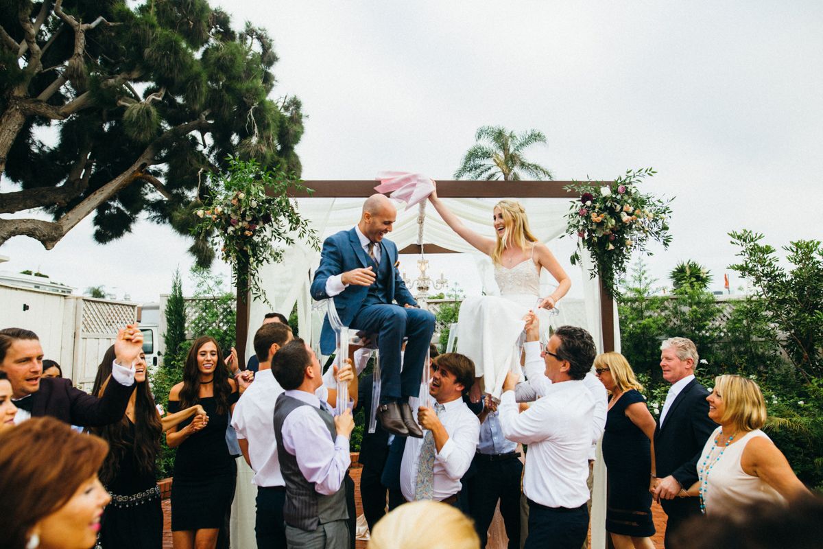 Bride and groom lifted by guests in chairs during a lively wedding celebration outdoors.