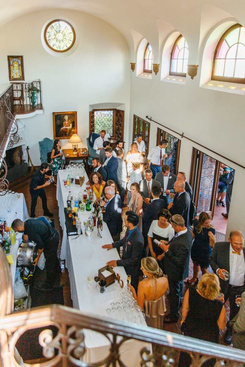 Aerial view of wedding cocktail hour guests mingling inside an elegant venue with arched windows and stained glass.