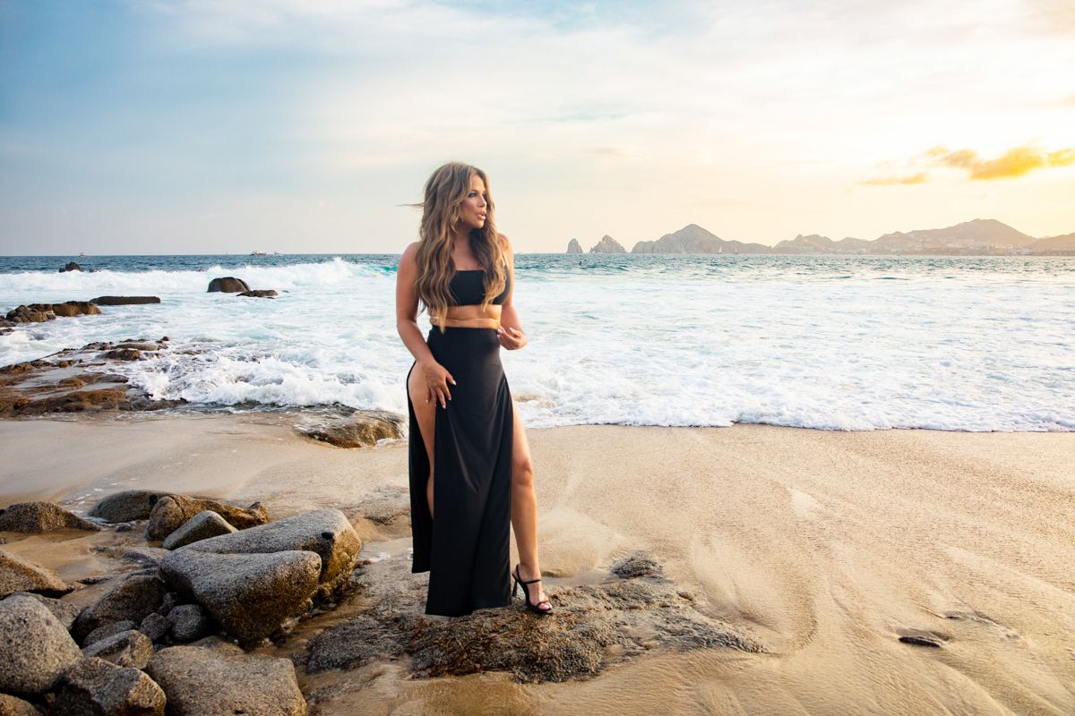 Woman in black two-piece outfit standing on rocky beach at sunset with ocean waves and cliffs behind.
