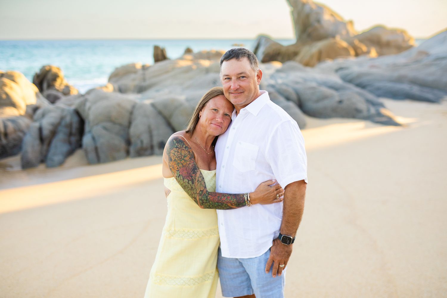 Couple embracing on a sandy beach with rocky backdrop, woman in yellow dress with tattoos, man in white shirt in cabo