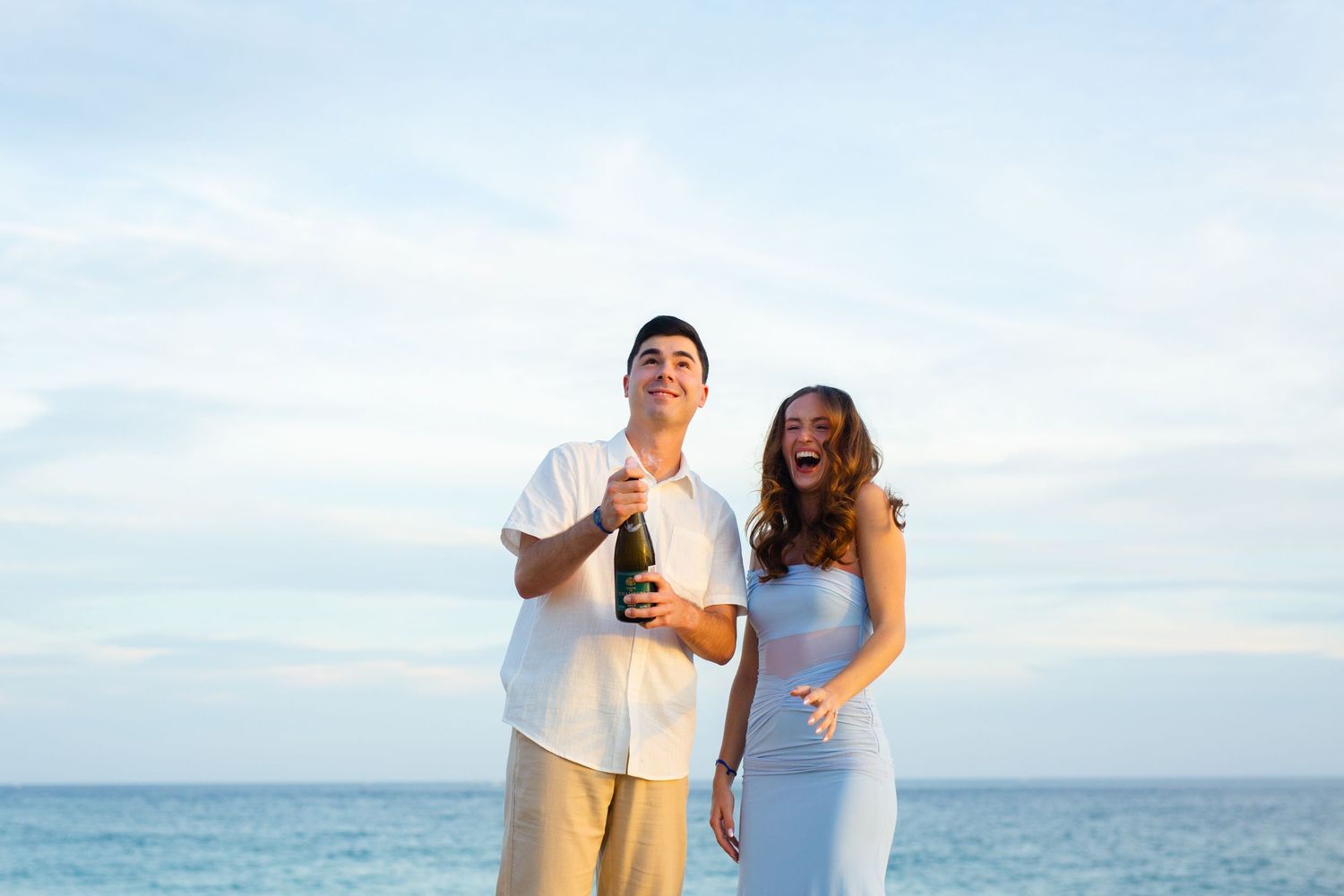 Couple celebrating on a beach, man holding champagne bottle, woman in blue dress, ocean backdrop in cabo