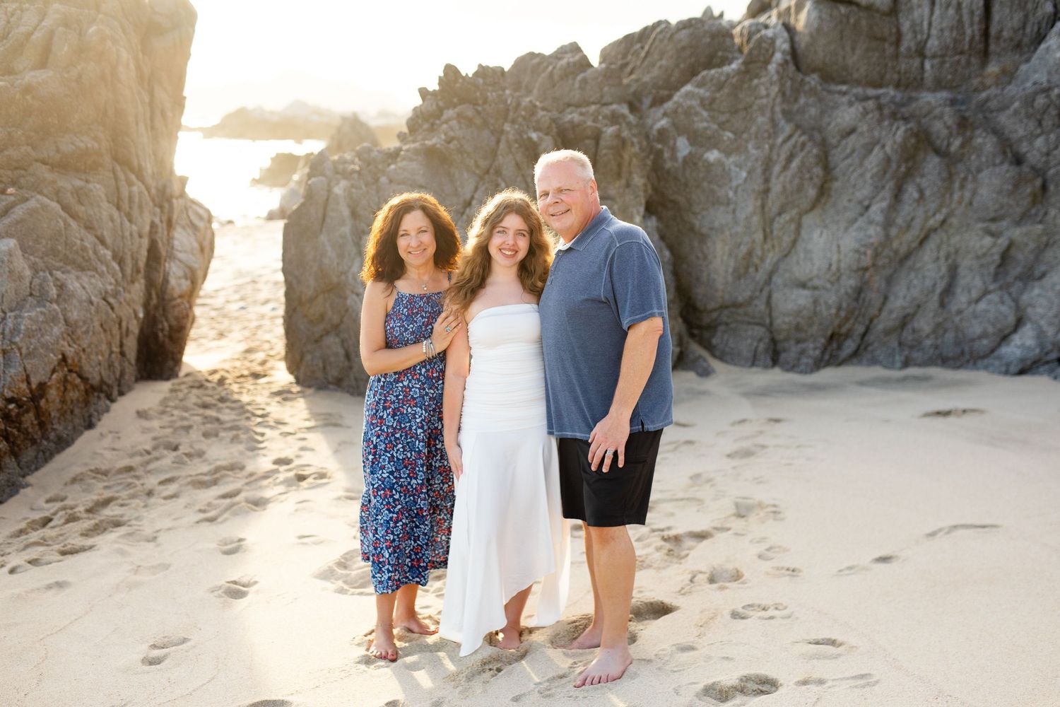 Three people smiling together on a sandy beach with rocky cliffs, wearing casual summer outfits in warm golden light in Cabo San Lucas.