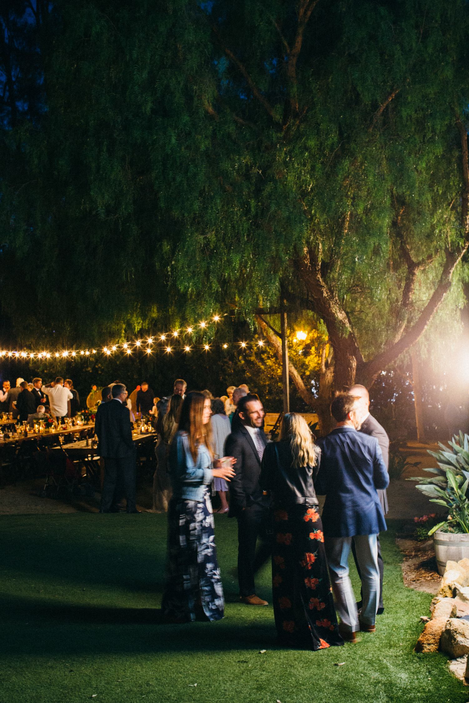 Guests mingle under string lights and trees at an elegant outdoor evening wedding reception.