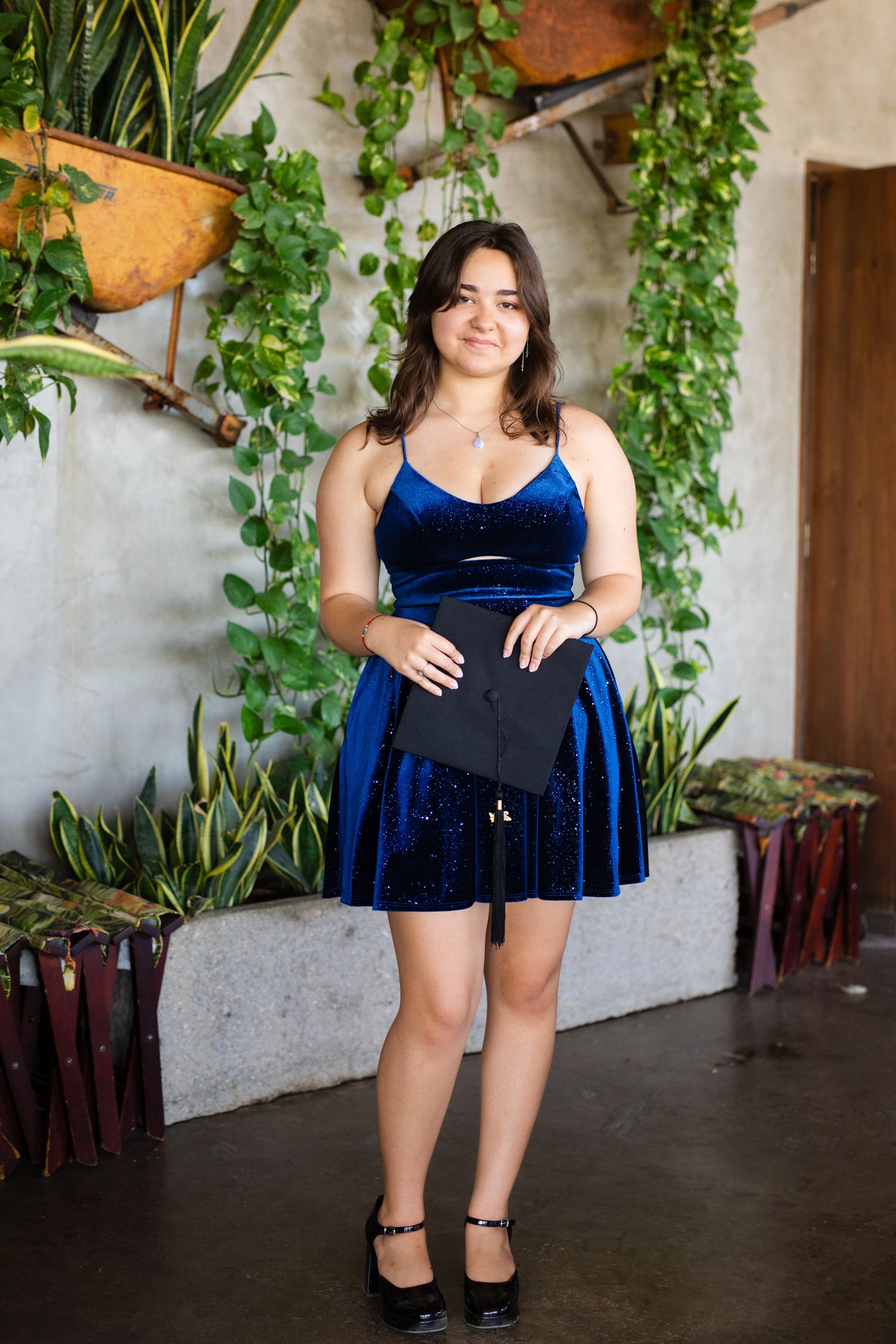 Young woman in blue velvet graduation dress holding a cap among lush green plants indoors.