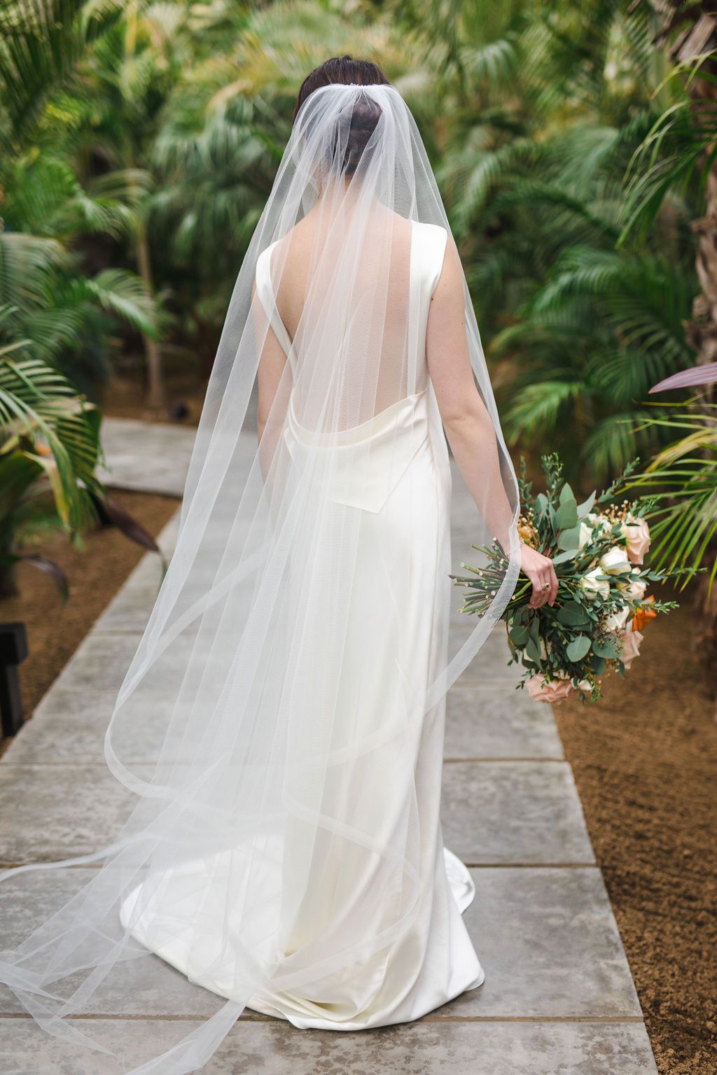 Bride in white gown and long veil walks along garden path holding floral bouquet, view from behind.