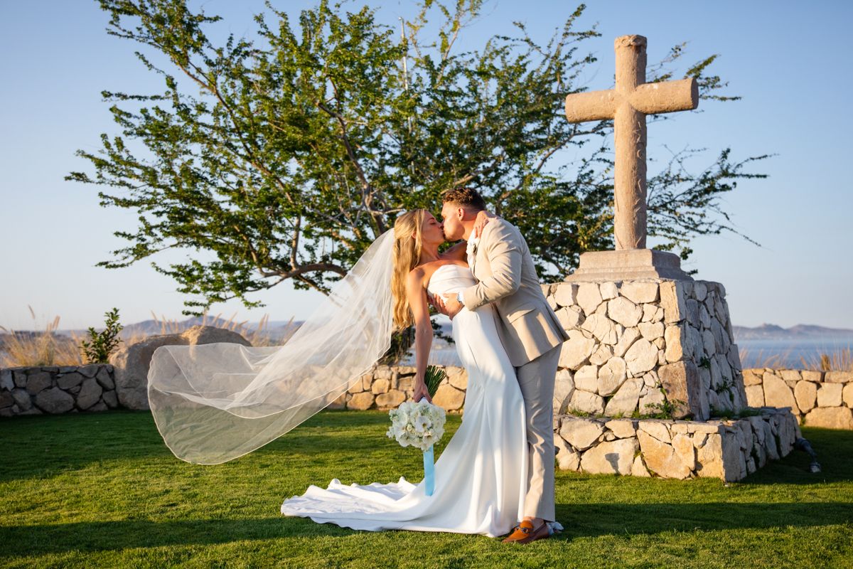 Bride and groom share first kiss beside stone cross at scenic outdoor wedding venue.
