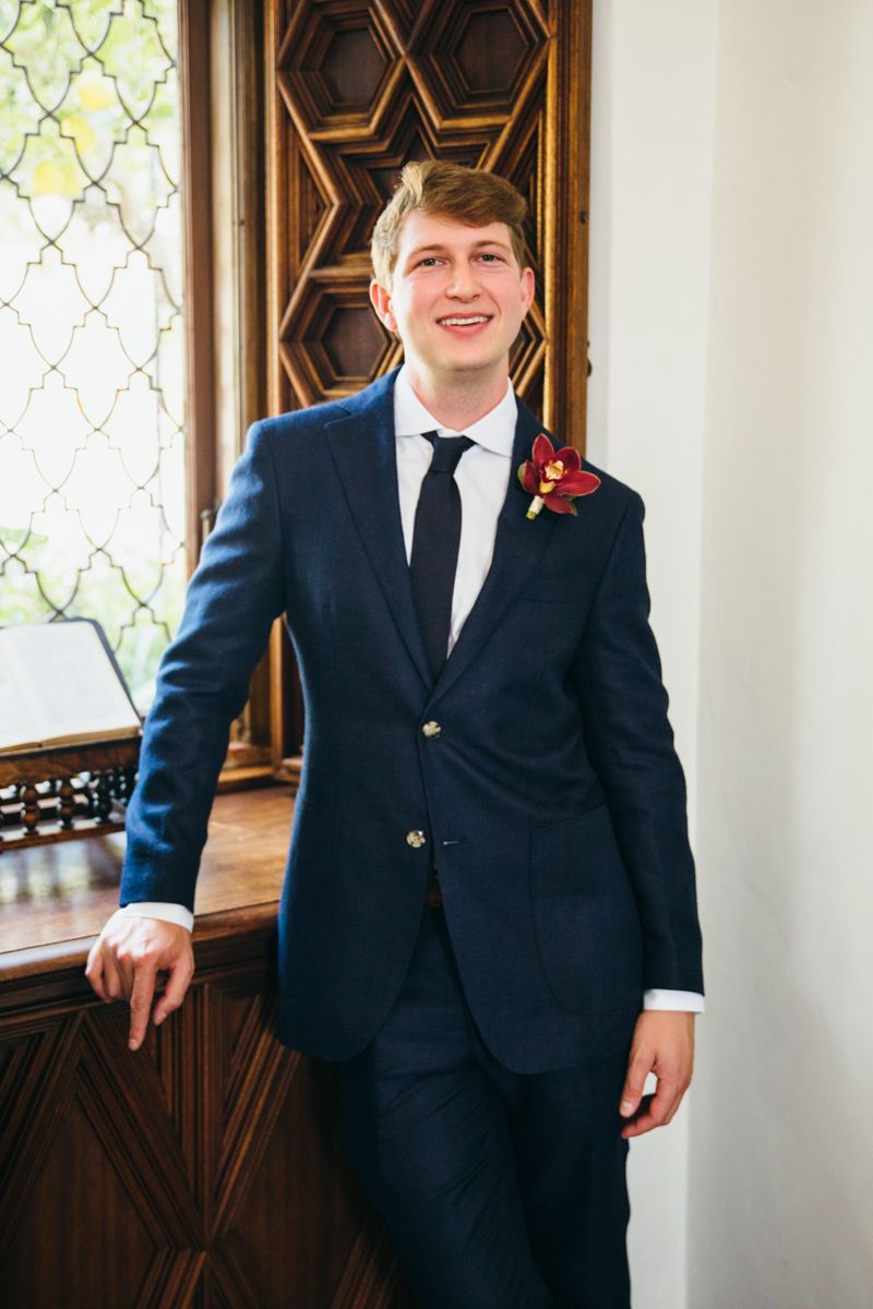 Smiling groom in navy blue suit with floral boutonniere leaning against a wooden window frame.