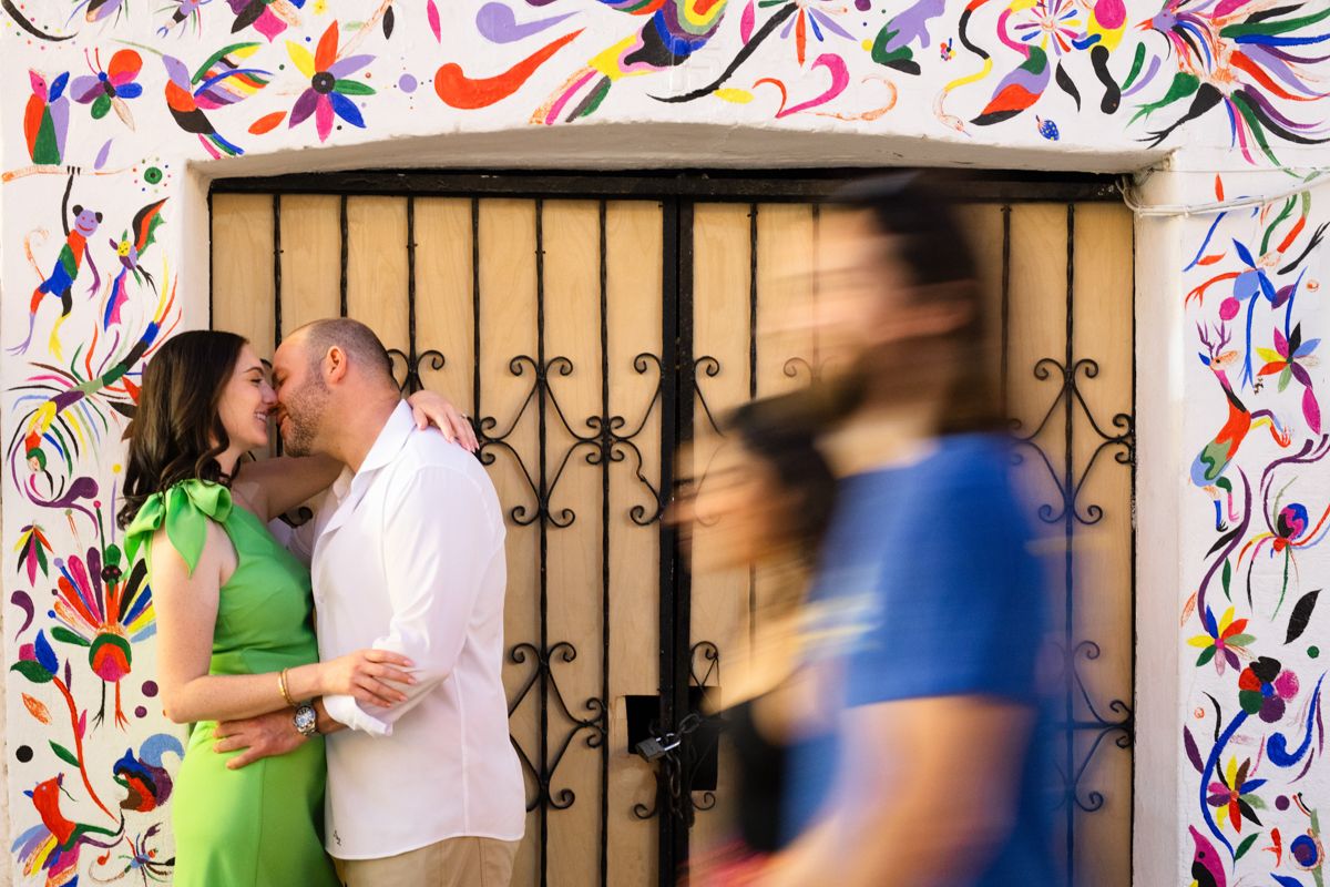 Couple kissing in front of vibrant otomi art mural gate with blurred passersby.