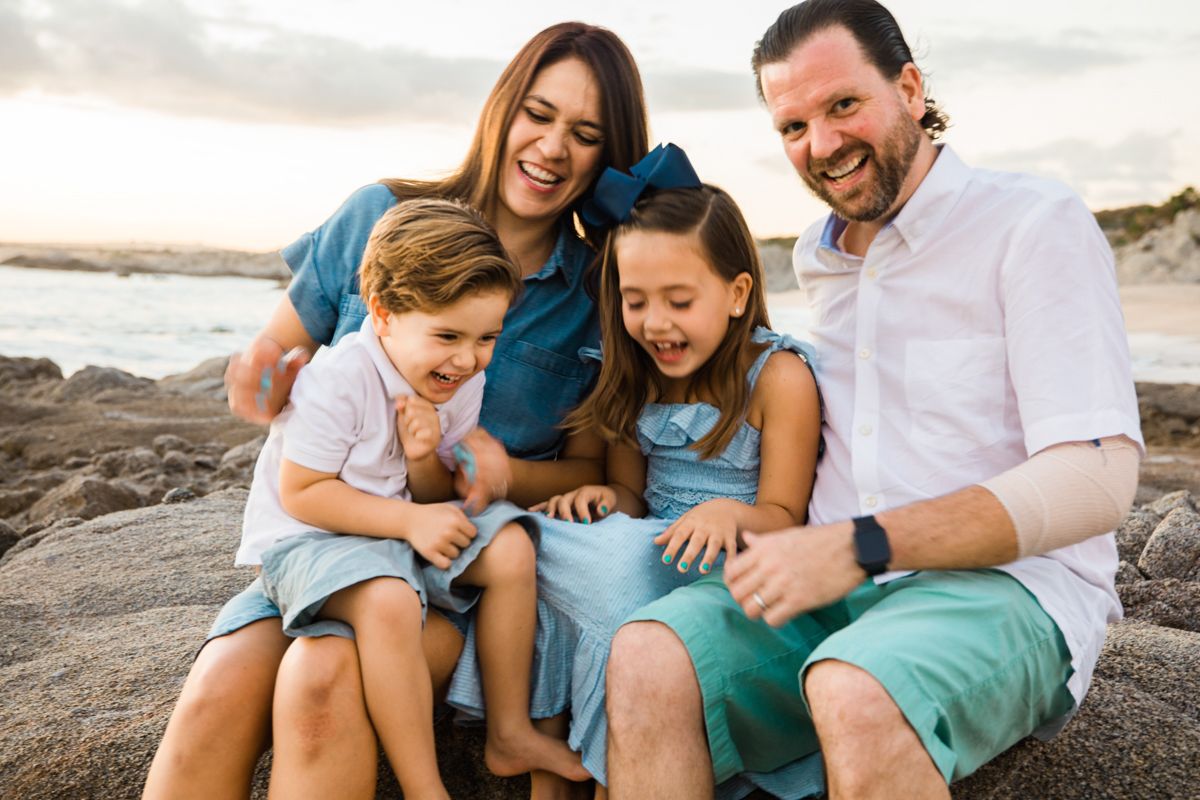 Happy family of four laughs together while sitting on rocks at a beach during sunset.