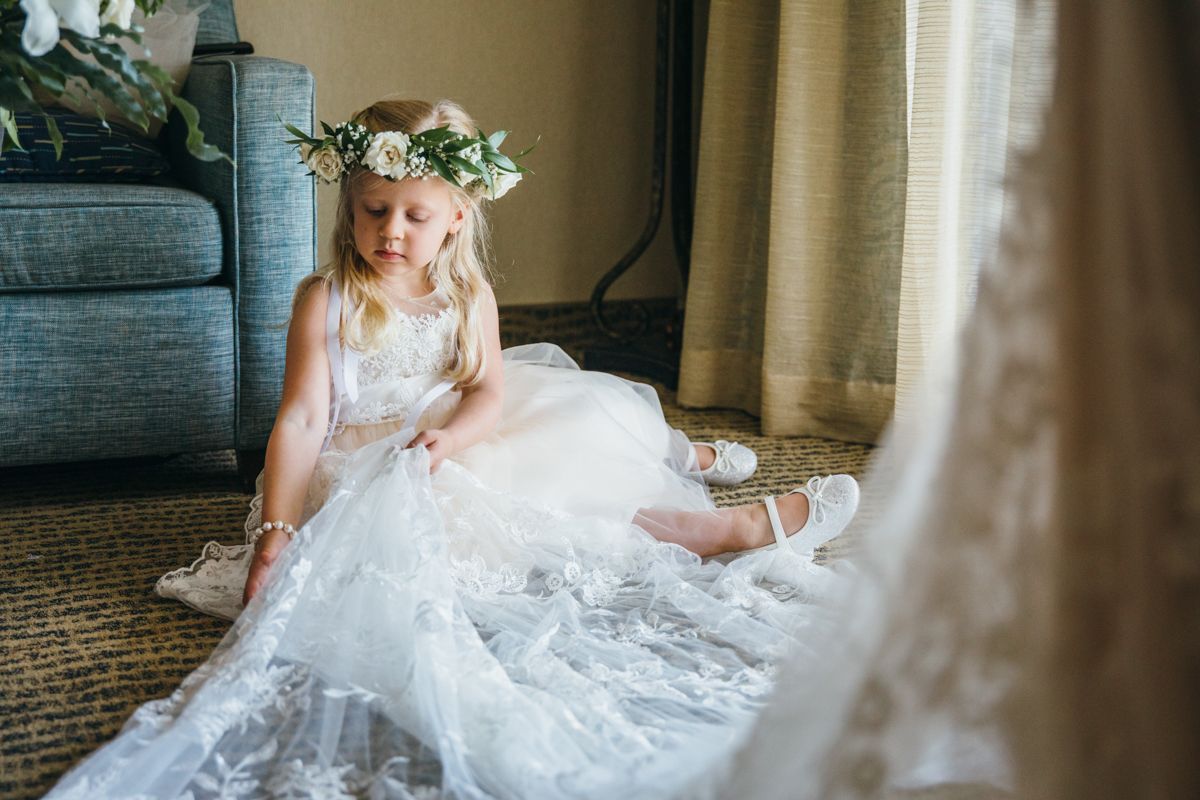 Young flower girl in white lace dress and floral crown sits on floor at wedding venue.