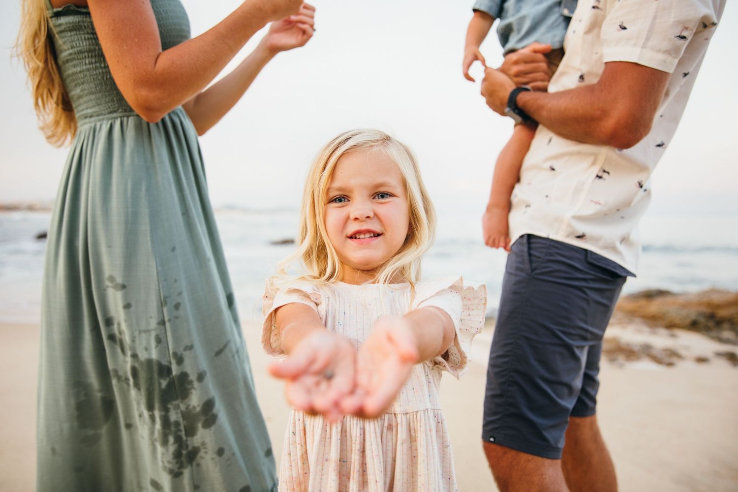 A smiling blonde girl in a white dress holds out her hands on a beach with her family behind her.