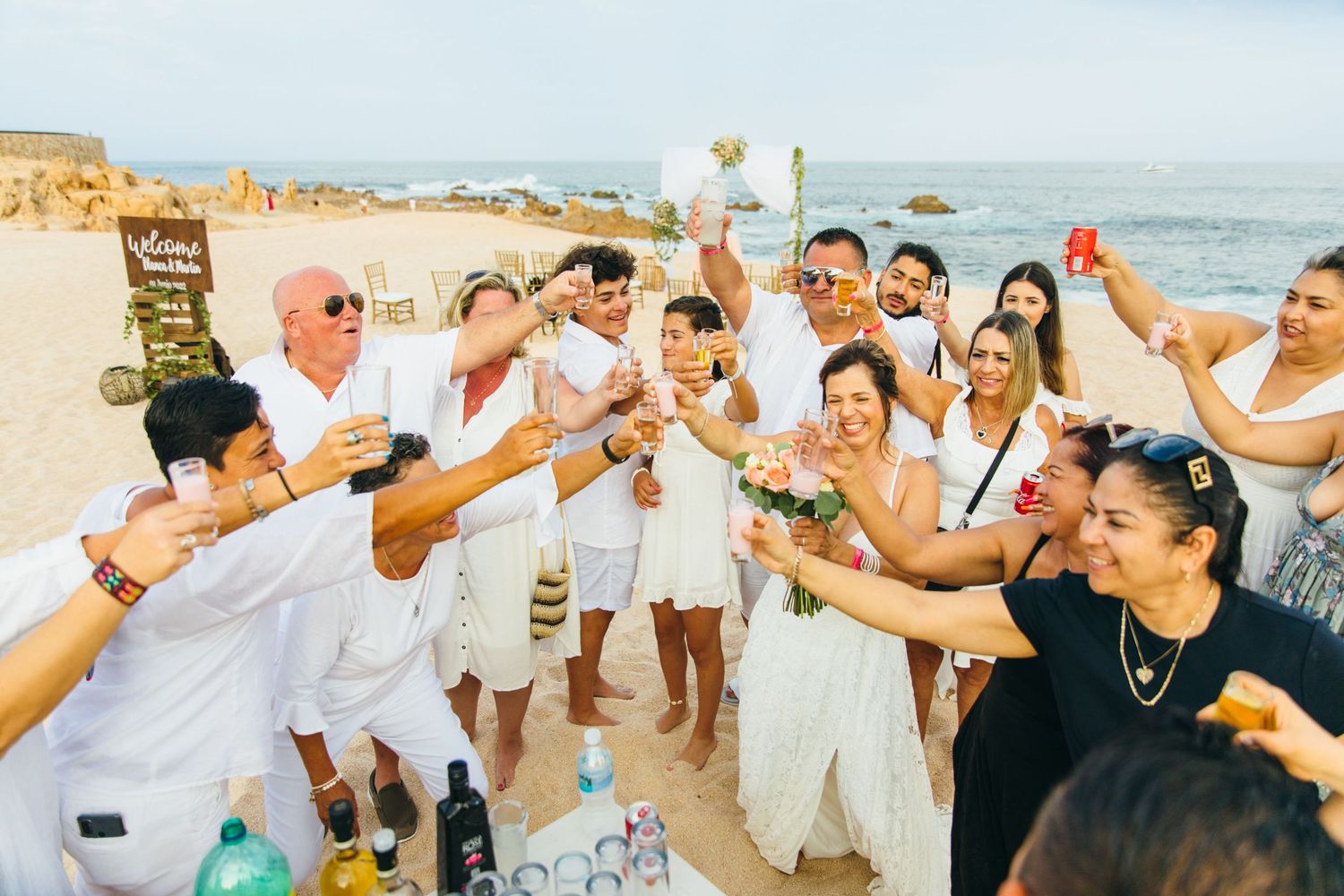Group of guests in white attire toasting at a cabo beach wedding celebration with ocean backdrop.