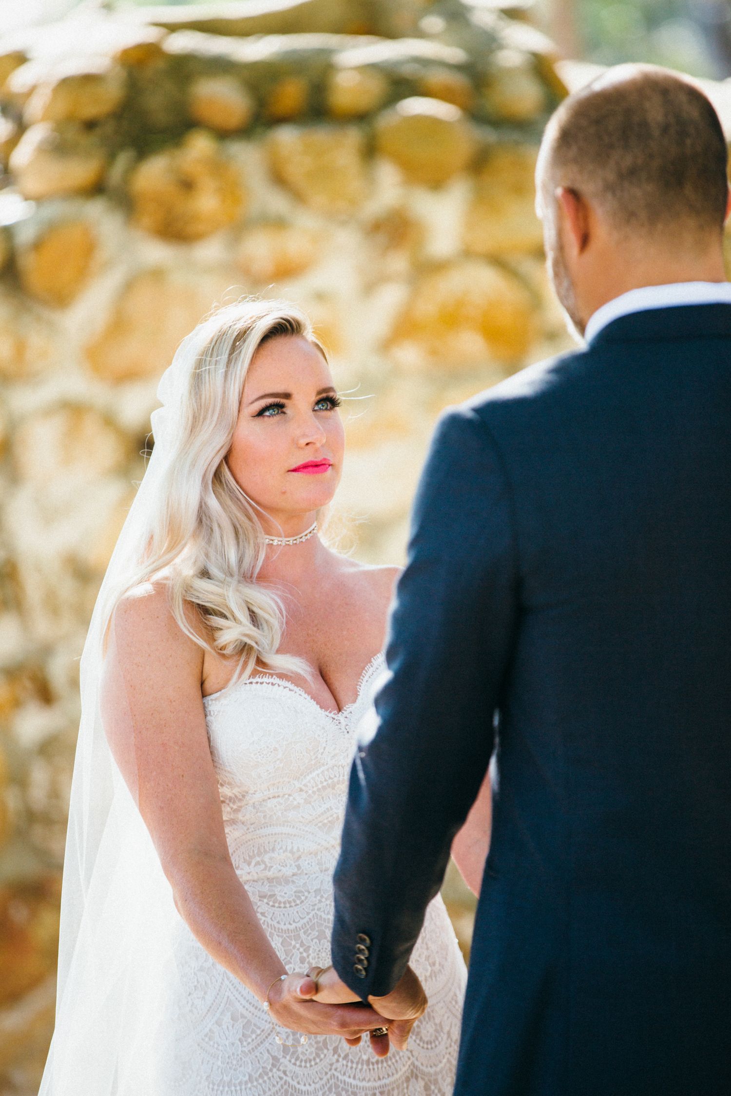 Bride in lace gown holds grooms hands during outdoor wedding ceremony near stone wall at Leo Carrillo ranch wedding