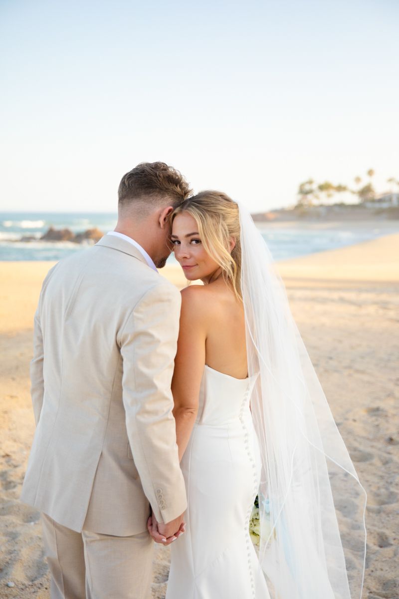 Bride and groom holding hands on a sandy beach at sunset, bride looking back.