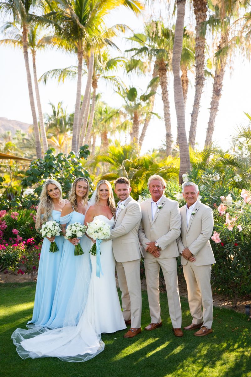Wedding party posing in tropical garden with palm trees, bridesmaids in light blue gowns.