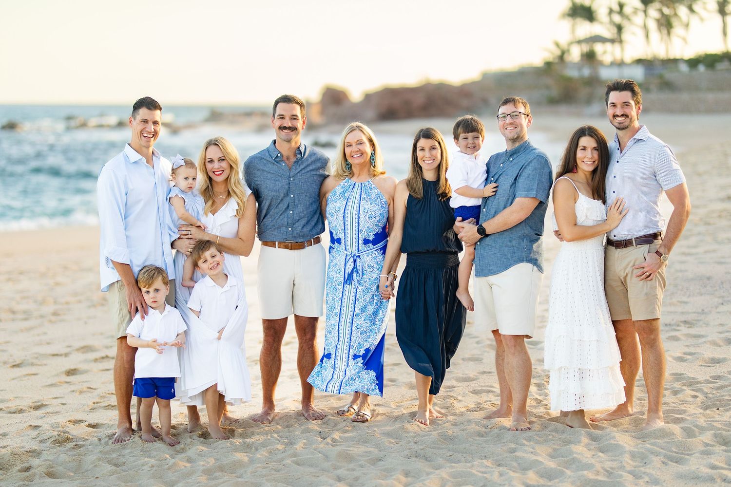 Large extended family of eleven posing together on sandy beach at golden hour.