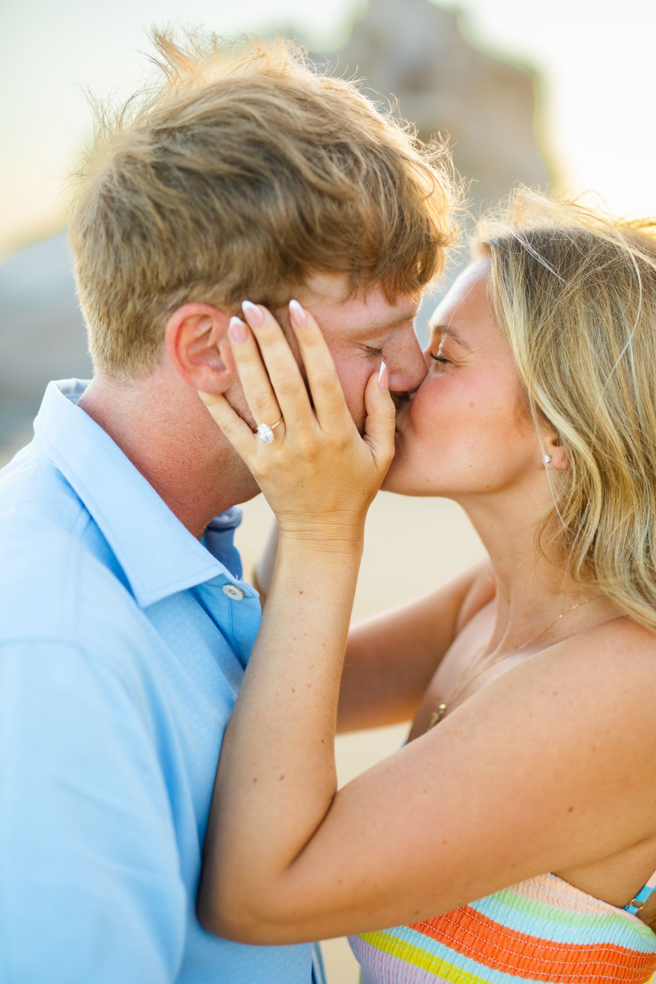 A couple shares a romantic kiss outdoors, with the woman showing off her engagement ring while embracing her partner.