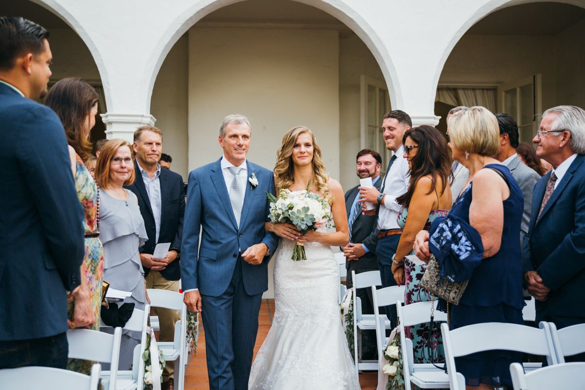 Bride walks down the aisle with her father past seated guests during an outdoor wedding ceremony.