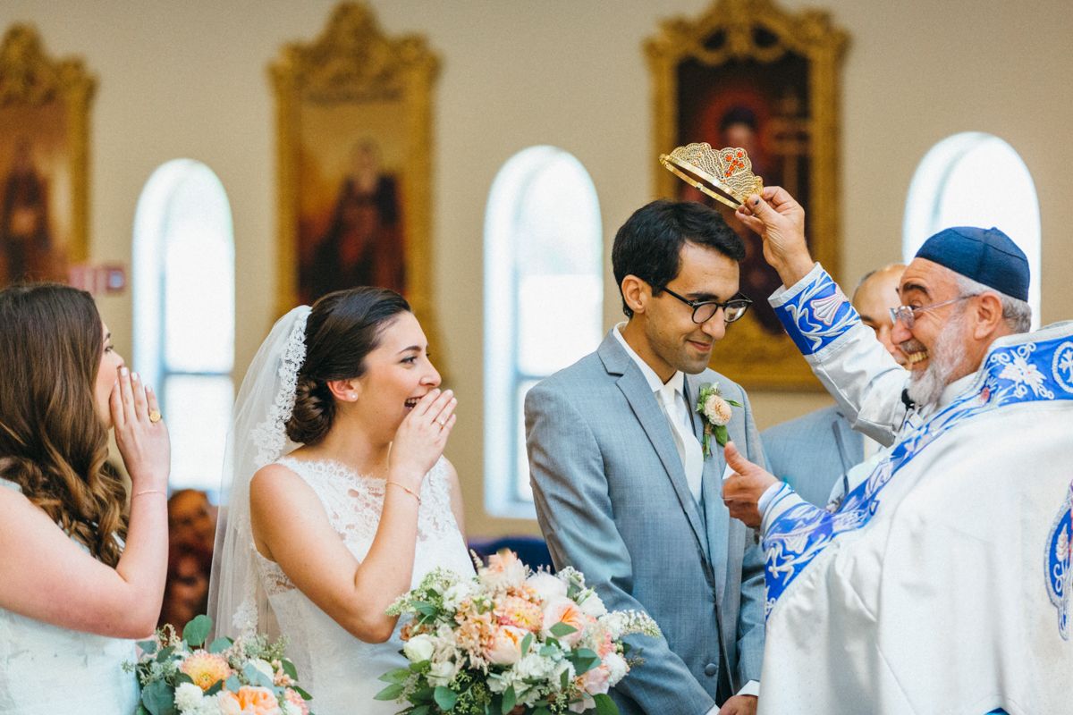 A bride laughs joyfully as an Orthodox priest places a crown on the groom during a wedding ceremony.