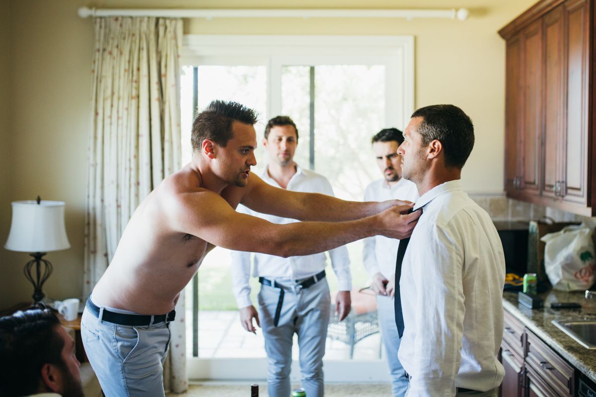 Shirtless groomsman helping groom fix tie while others watch during wedding preparations.