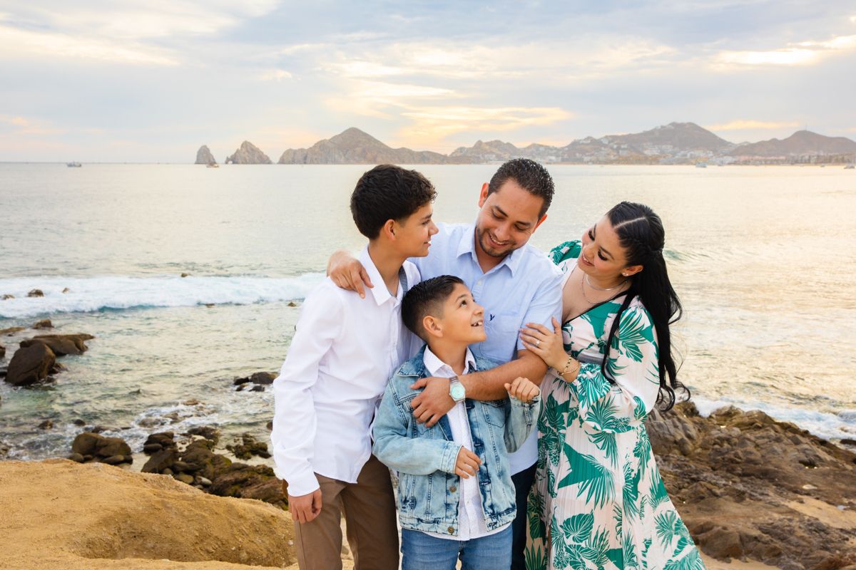 Happy family of four shares a warm embrace on rocky beach with scenic ocean sunset backdrop.