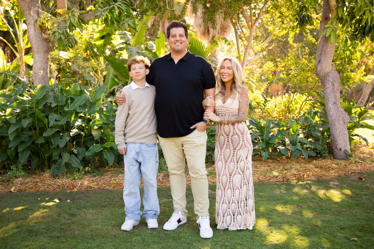 Family of three posing together on green lawn surrounded by lush garden trees.
