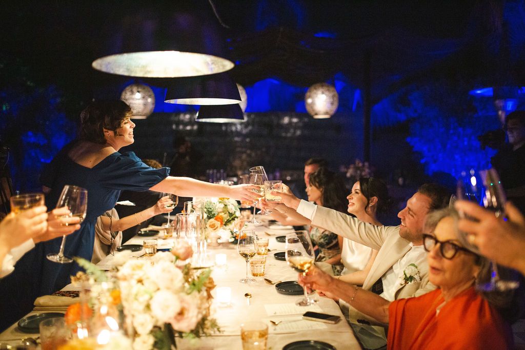 Wedding guests raising glasses in a toast at a candlelit dinner reception under warm pendant lights.