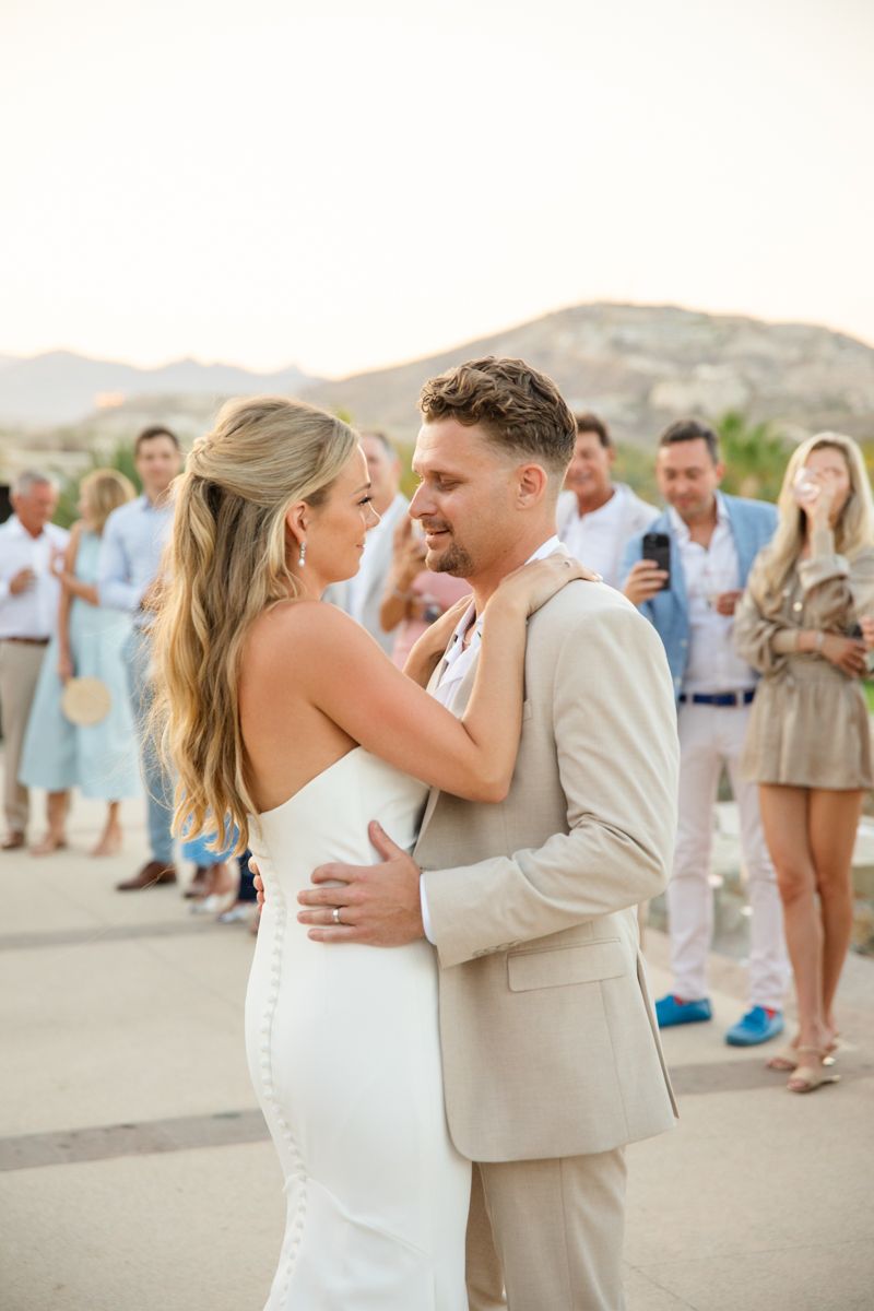 Bride and groom sharing first dance outdoors at sunset with guests watching.