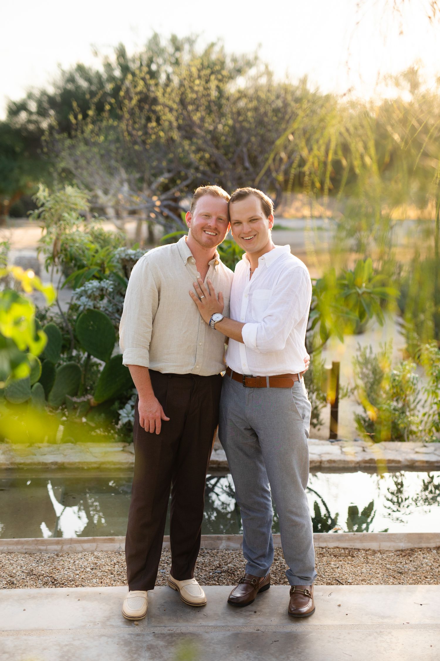 Two men smiling and embracing outdoors near a pond with lush greenery in warm golden hour light.
