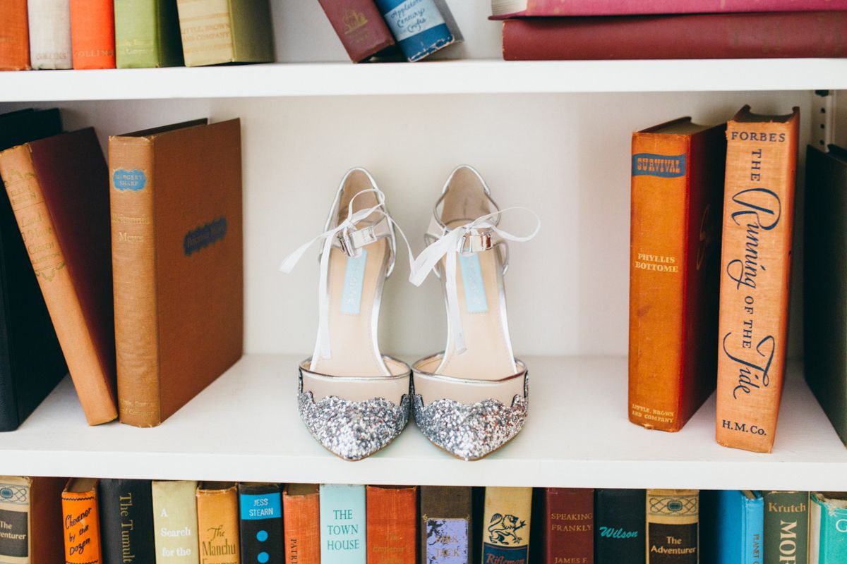 Glittery bridal heels displayed on a white bookshelf surrounded by colorful vintage books.