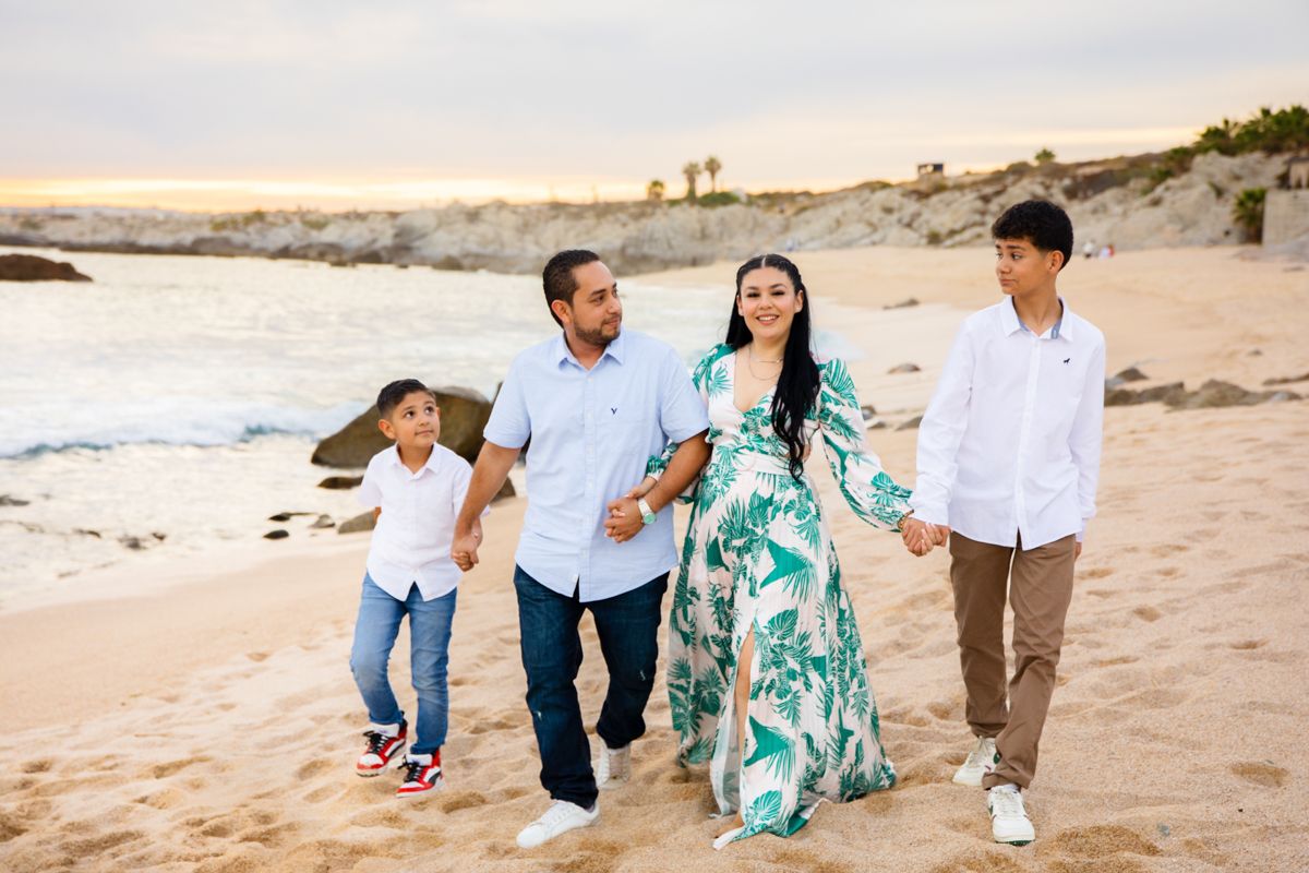 Family of four walking hand in hand on a sandy beach at sunset, wearing colorful outfits near rocky cliffs.