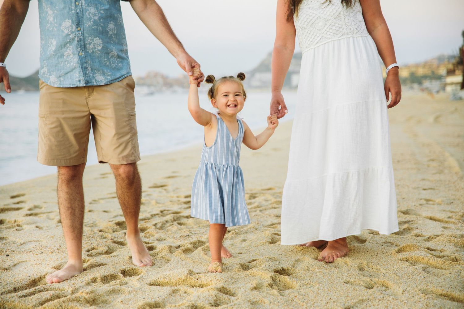A happy toddler girl in a blue dress holds hands with her parents while walking barefoot at Medano beach in Cabo San Lucas, Mexico.