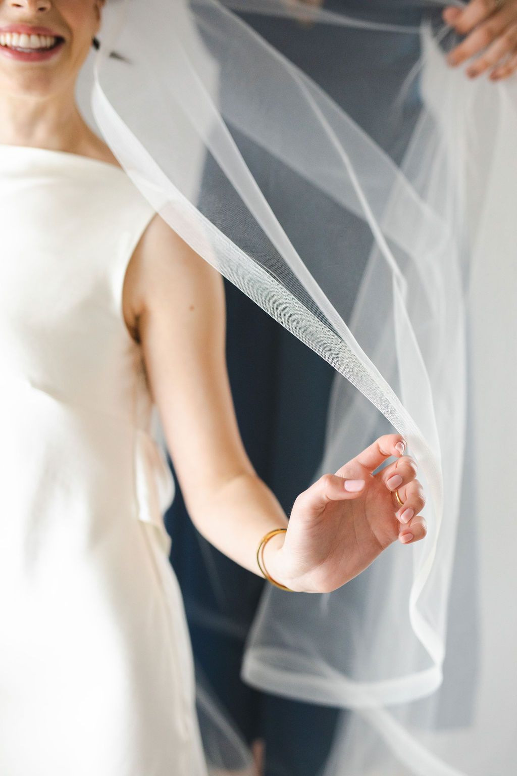 Smiling bride in sleeveless white wedding dress holding flowing tulle veil with gold bangle bracelet.