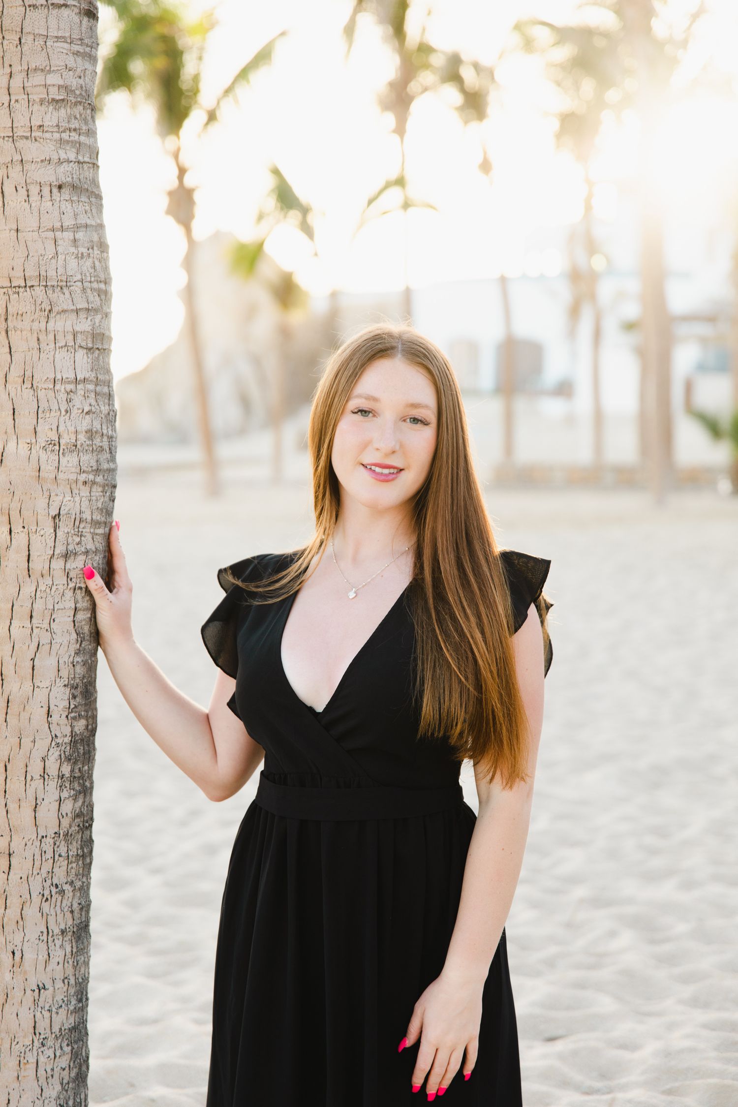 Redhead woman in black dress leaning against palm tree on sandy beach at golden hour.