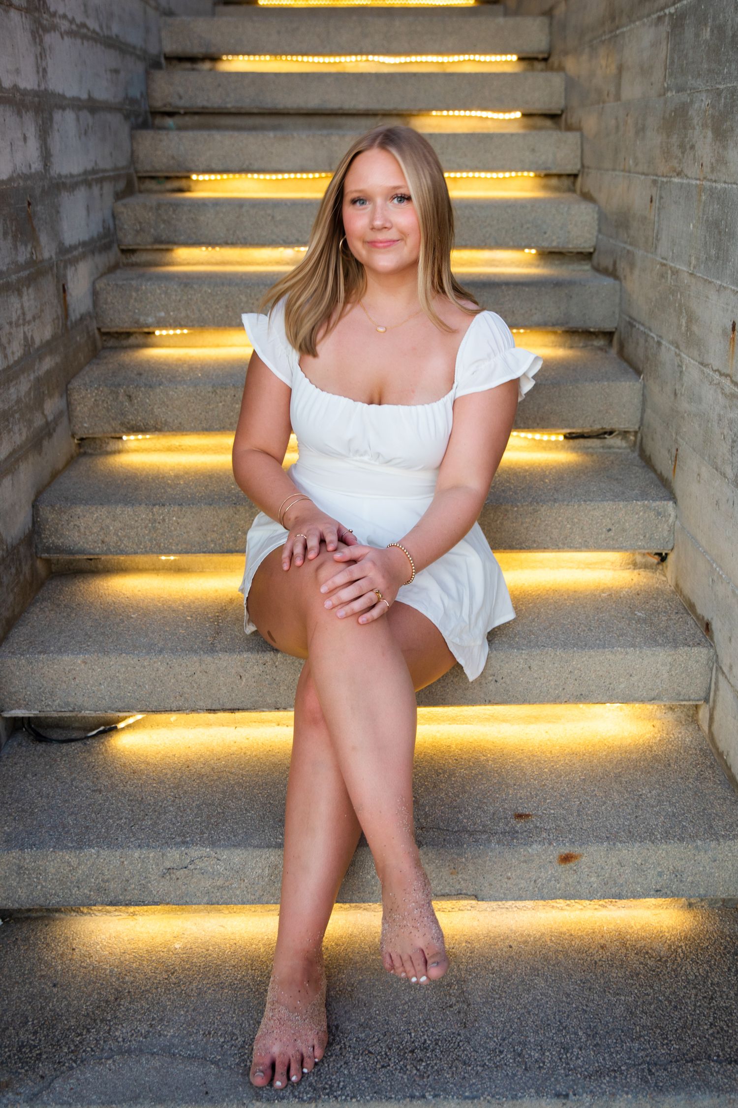 Blonde woman in white dress sitting barefoot on illuminated stone steps with golden LED lighting.