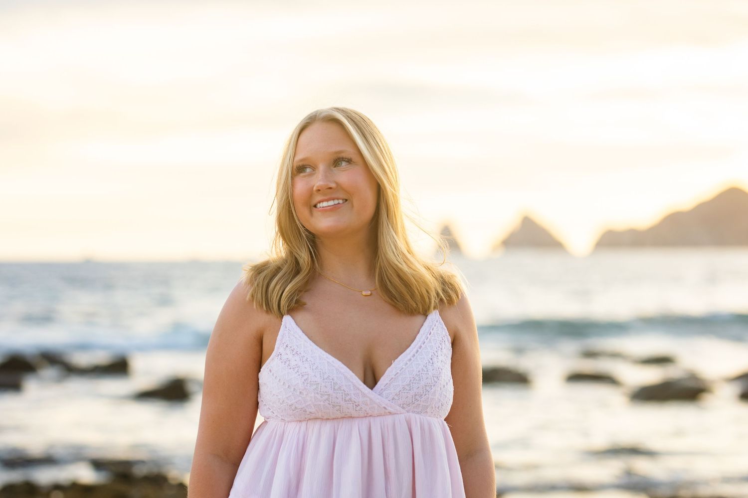 Smiling blonde woman in pink dress standing on a rocky beach at golden hour sunset.