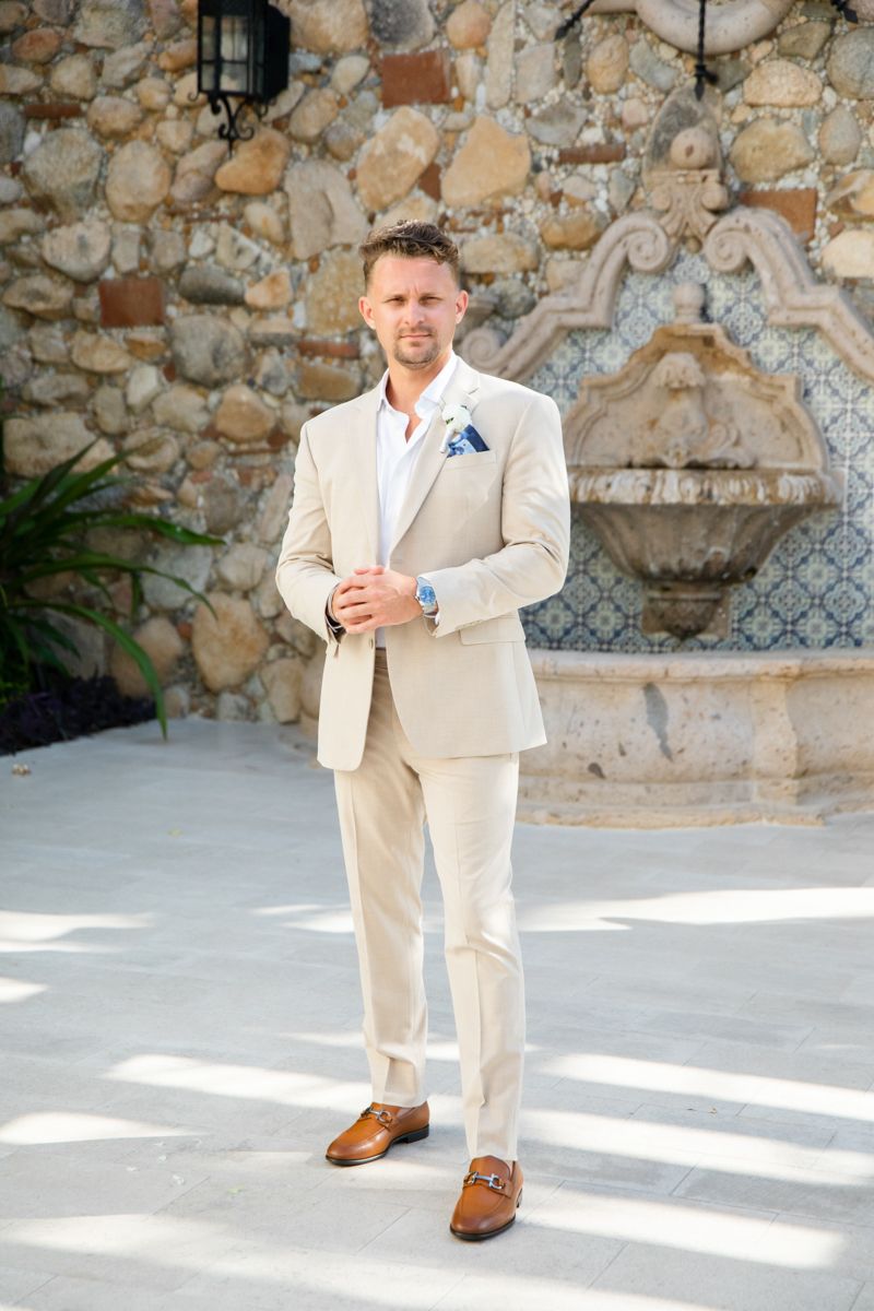 Groom in beige suit with blue pocket square stands before stone wall with fountain.
