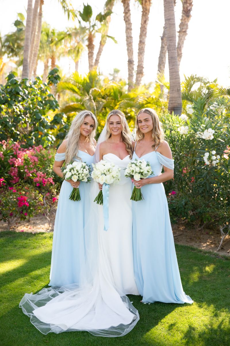 Bride flanked by two bridesmaids in light blue gowns holding white bouquets outdoors.