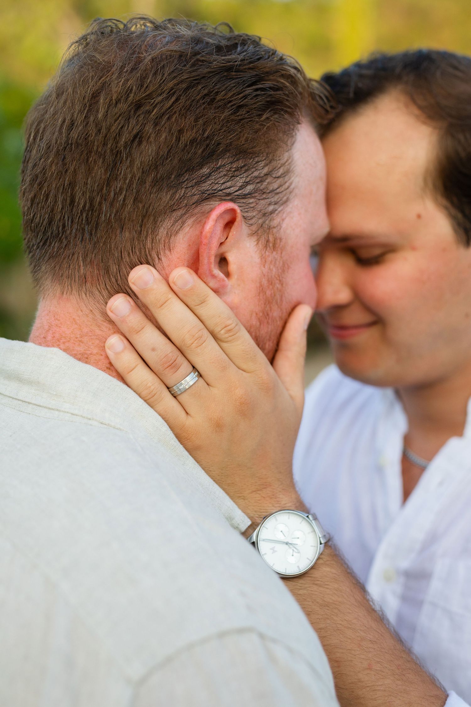 Close-up of same-sex couple touching foreheads, one wearing an engagement ring and watch.