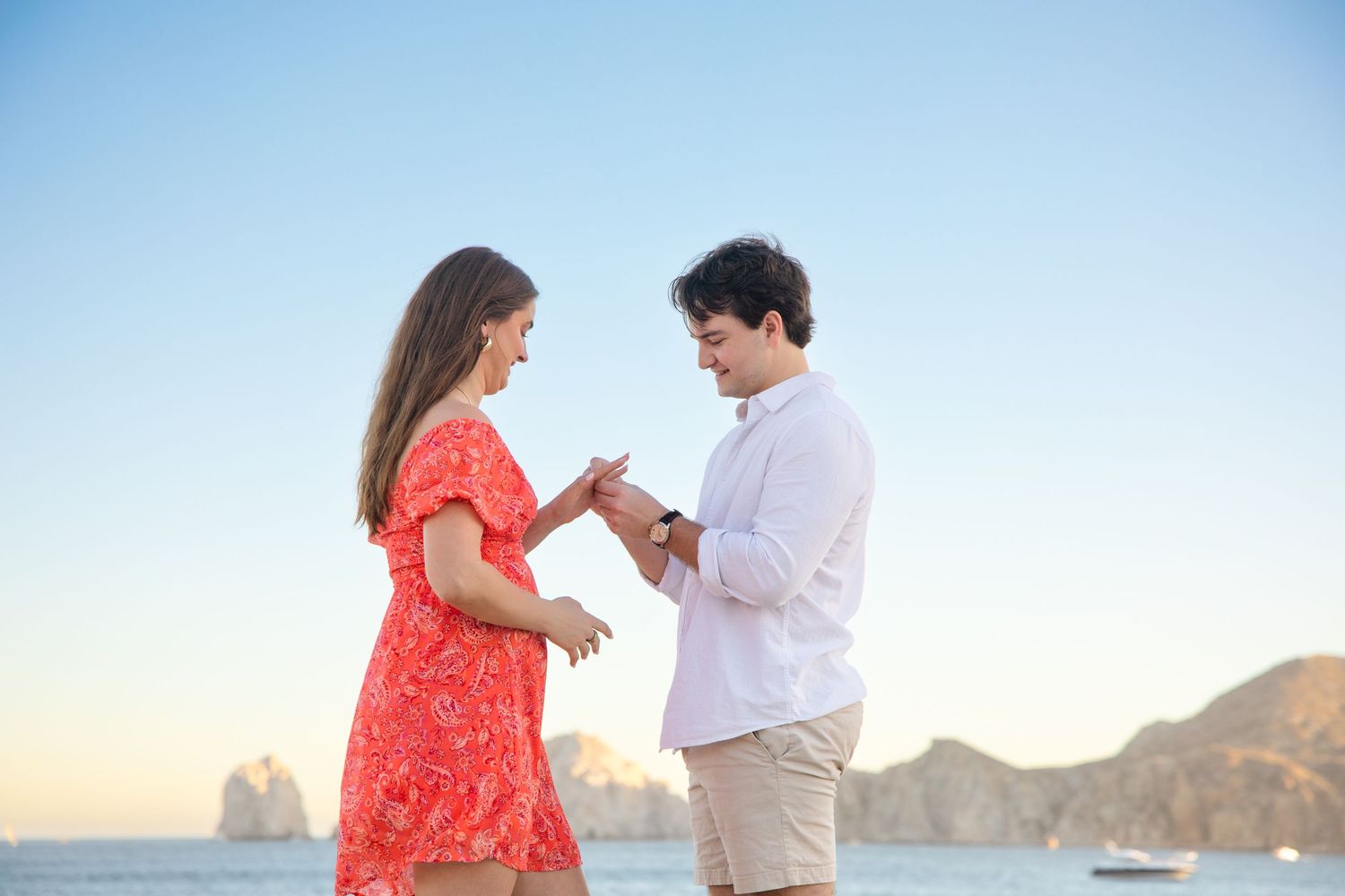 Man proposing to woman in red dress on a beach with Cabo San Lucas arch visible in background.
