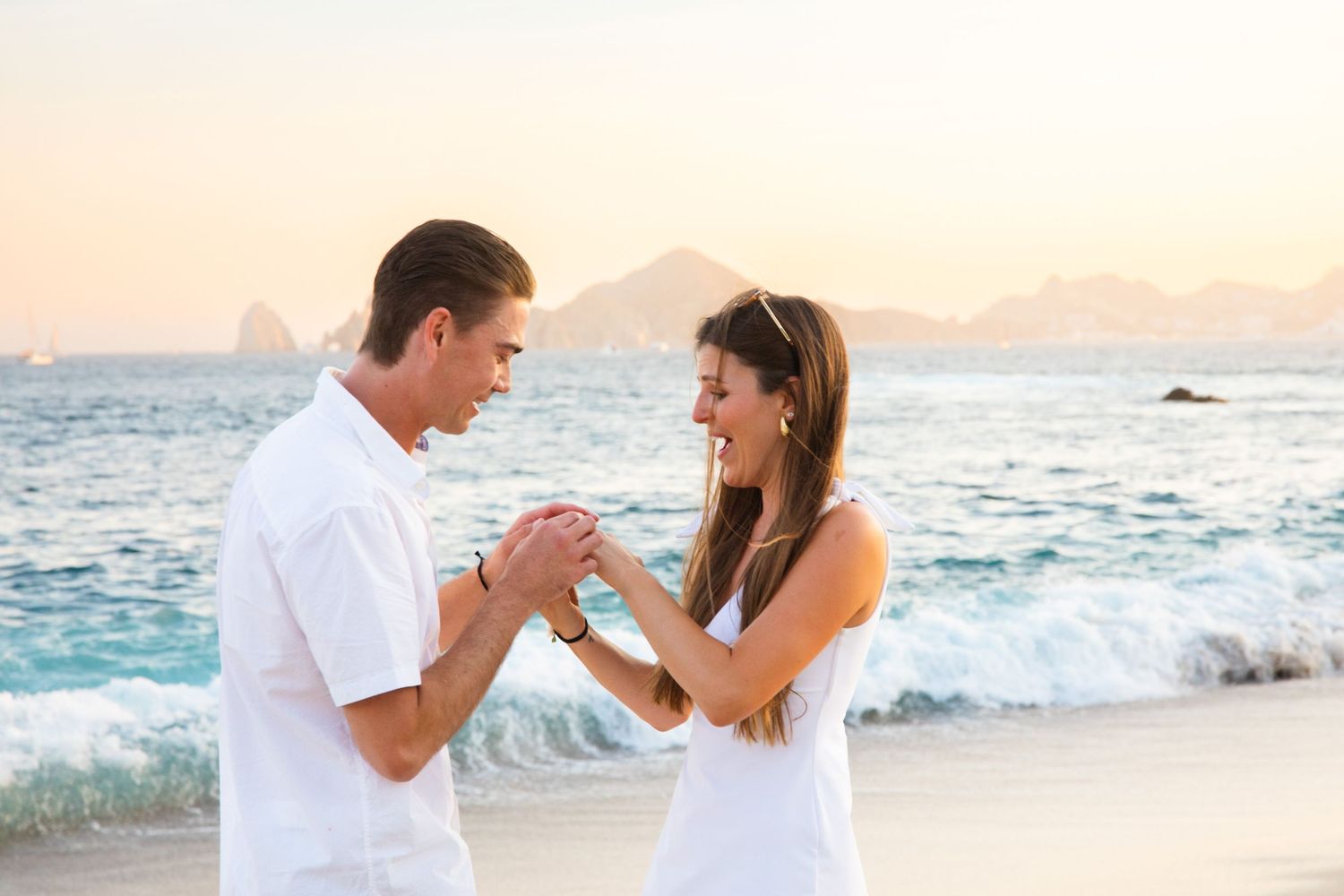 Happy couple holding hands on a beach at sunset, both dressed in white.