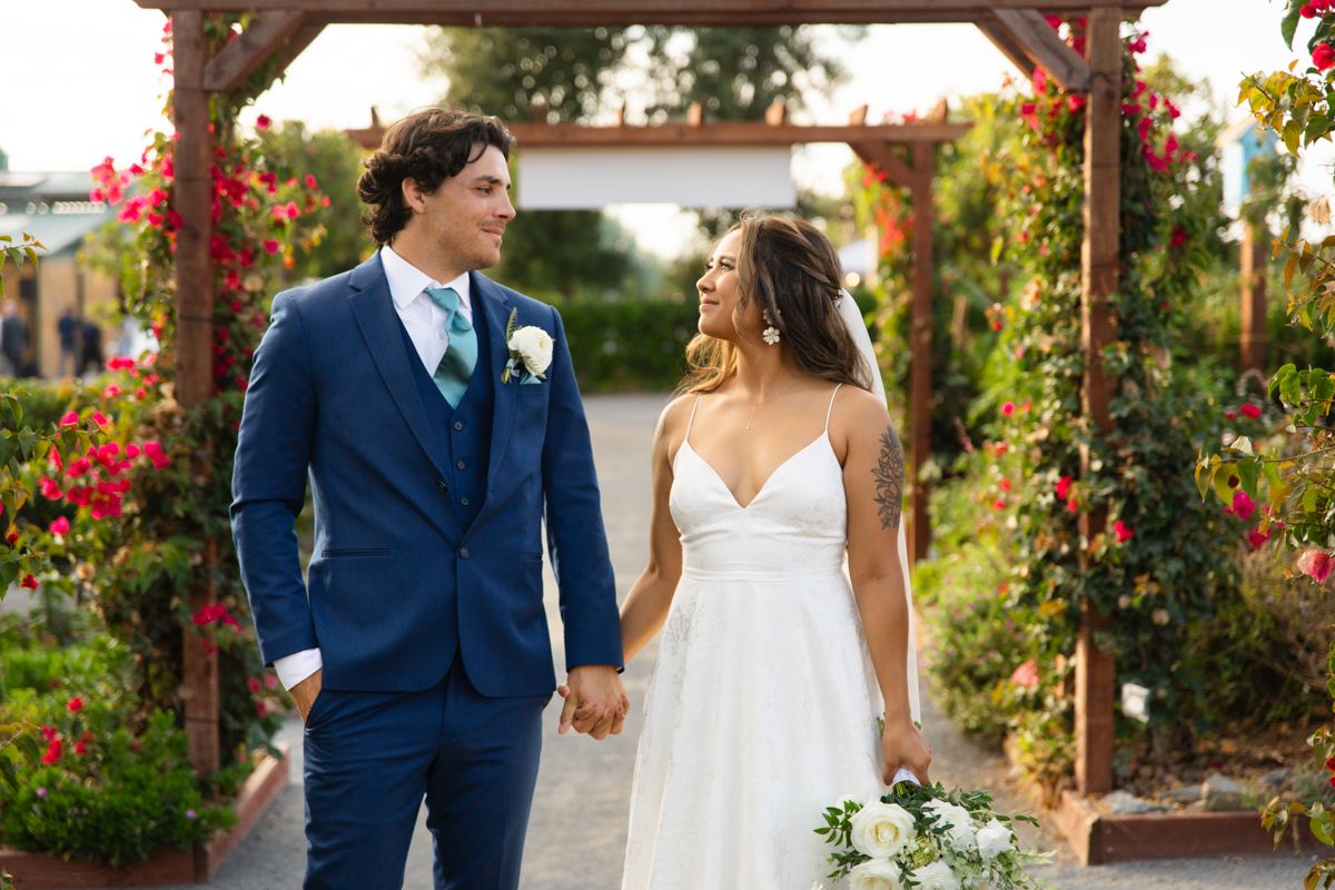 Bride and groom holding hands walking through a garden arbor with colorful flowers, groom in navy suit.